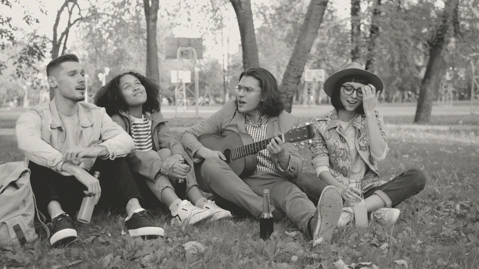 Four friends sitting on the grass in a park, one playing guitar.